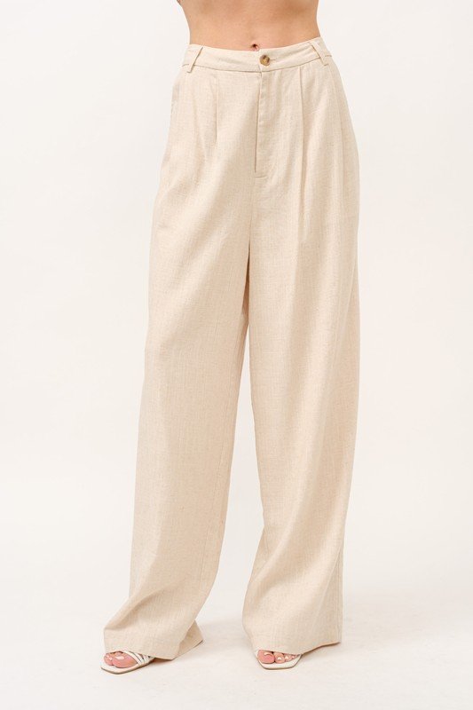 A woman stands facing forward, modeling the And The Why high-rise wide leg linen blend pants in beige, paired with a simple black top.